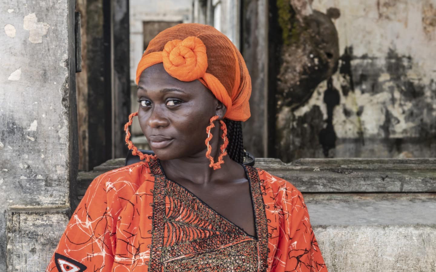 A Black Woman Poses For The Camera. She Is Wearing An Orange Dress With An Orange Headwrap And Orange Earrings.