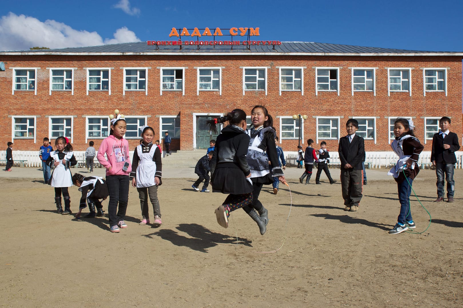 Mongolian Children Playing Jump Rope In Front Of School Building.