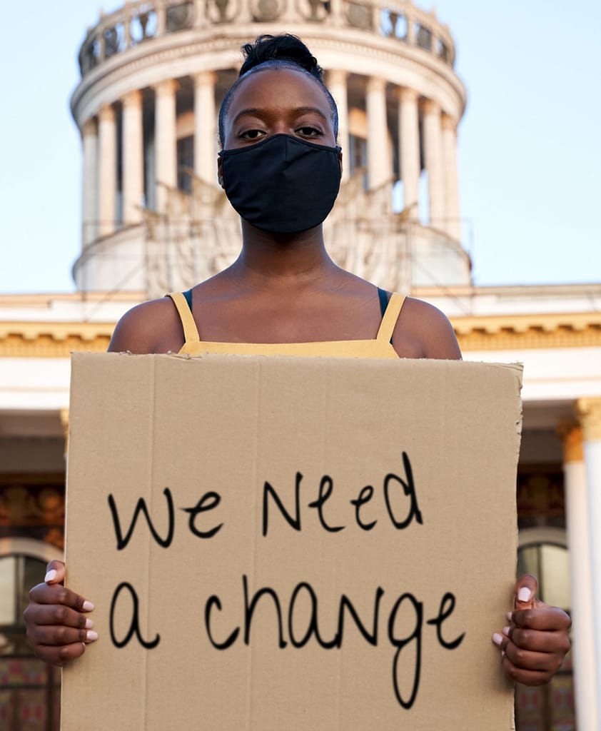 Black woman wearing mouth mask holding a placard saying we need a change