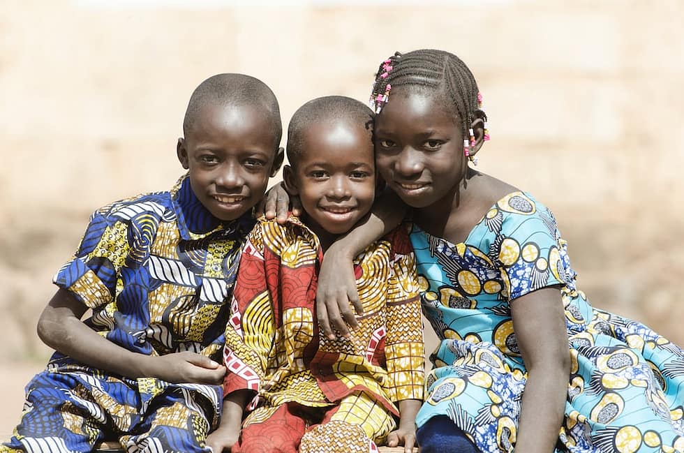 Three children wearing African print with their arms around each other's shoulders smiling at the camera