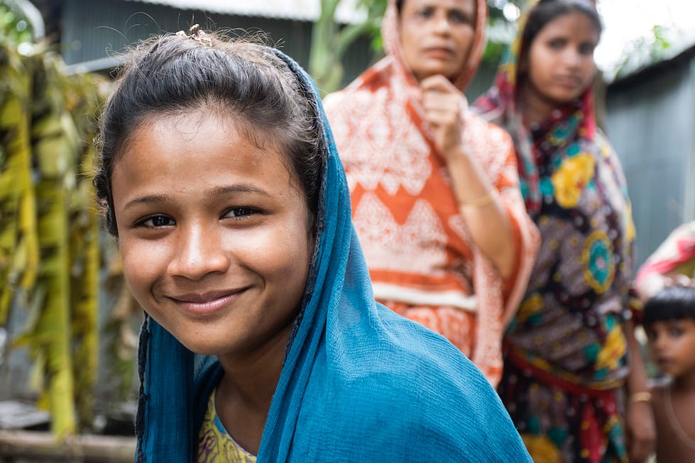 Bangladeshi girl wearing a headscarf smiling at camera, two women and a boy stand behind her