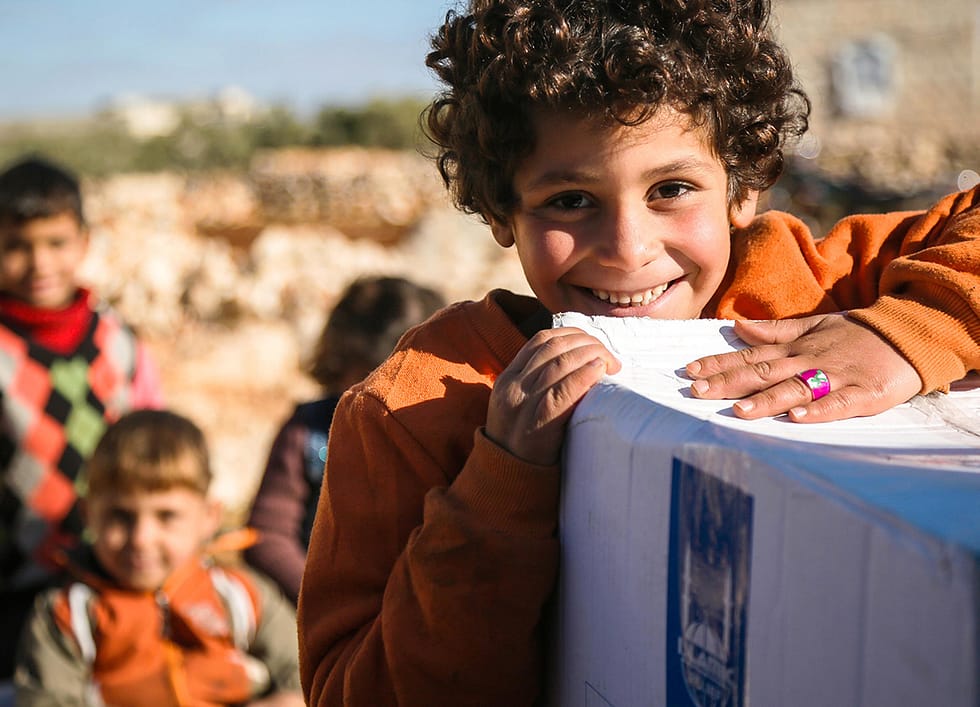 Arabic boy smiles at the camera with his hands resting on a Islamic Relief box