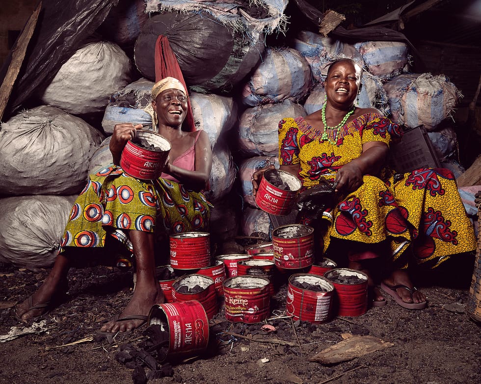 Two black women wearing African print selling cans of charcoal sat in front of large bags of charcoal in Togo
